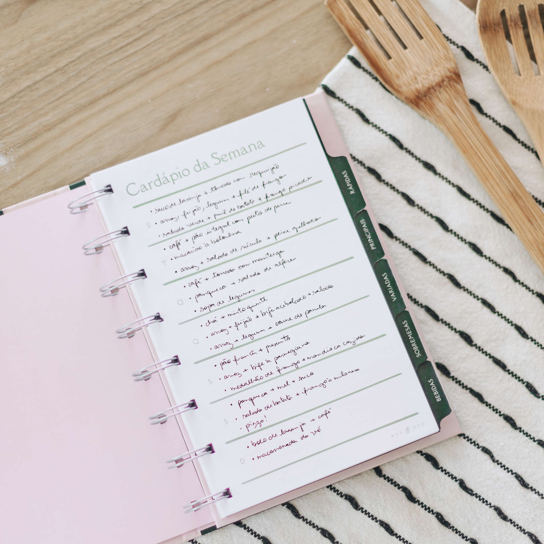 caderno de receitas com cardápio da semana sobre fundo de madeira com pano de prato e colheres de madeira ao lado