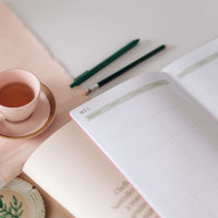 caderno planejador mensal sobre mesa. ao fundo vemos uma caneta, um lápis e uma xícara de chá